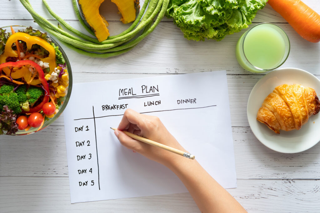 calories control, meal plan, food diet and weight loss concept. top view of hand filling meal plan on weekly table with salad and fresh vegetable on dining table